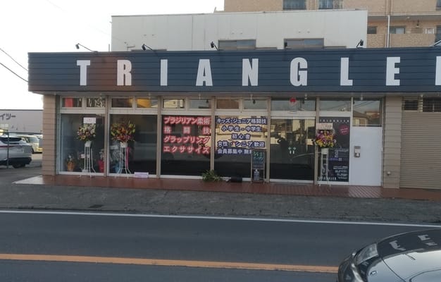 ジムの外観で、「TRIANGLER」の大きな看板が目立ち、初心者から女性、子どもまで歓迎する内容が窓に表示されています。